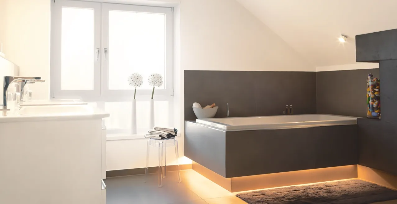 Moderne Eckbadewanne mit indirekter LED-Beleuchtung in hellem Dachbad mit minimalistischer Einrichtung und großen Fenstern.
