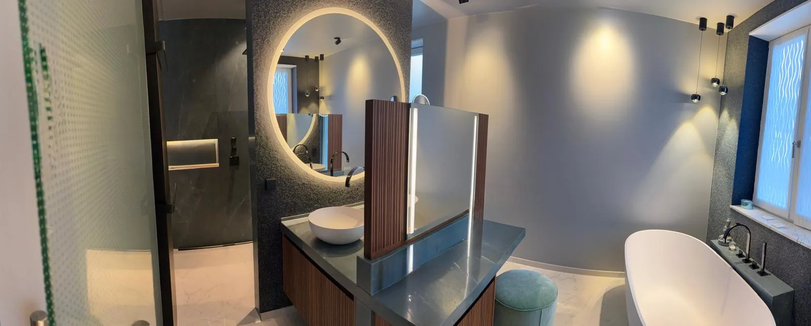 Modernes Bad mit freistehender Badewanne, beleuchtetem Rundspiegel, Natursteinoberflächen und stilvollem Waschtisch aus Holz.