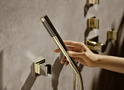 Goldene Handbrause der Serenity Sky von Dornbracht, wird gehalten mit glänzenden Armaturen und eleganten, minimalistischen Bedienelementen im Hintergrund.
