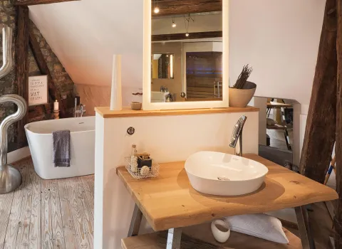 Stilvolles Badezimmer im Dachgeschoss mit freistehender Badewanne, Natursteinwand, Holzbalken und modernem Waschplatz aus Holz mit beleuchtetem Spiegel.