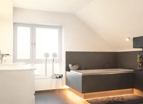 Moderne Eckbadewanne mit indirekter LED-Beleuchtung in hellem Dachbad mit minimalistischer Einrichtung und großen Fenstern.