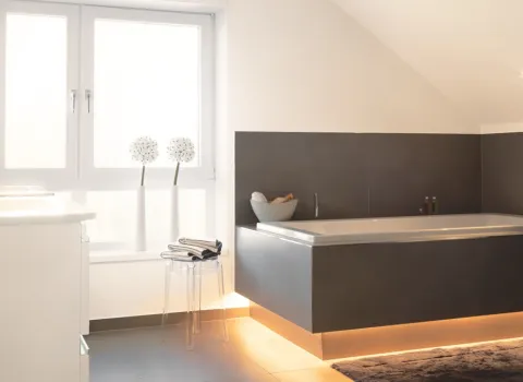 Moderne Eckbadewanne mit indirekter LED-Beleuchtung in hellem Dachbad mit minimalistischer Einrichtung und großen Fenstern.