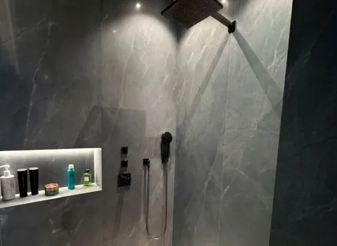 Moderne Walk-in-Dusche mit großformatiger Natursteinoptik, schwarzer Regendusche und integrierter beleuchteter Duschablage.