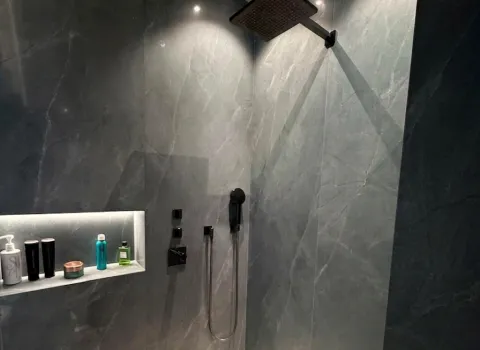Moderne Walk-in-Dusche mit großformatiger Natursteinoptik, schwarzer Regendusche und integrierter beleuchteter Duschablage.