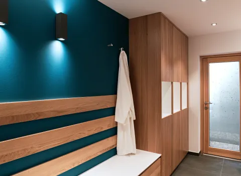 Moderner Wellness-Eingangsbereich mit dunkelblauer Akzentwand, Holzpaneelen, integrierter Sitzbank, großem Holzschrank und Bademantel an einem Haken.