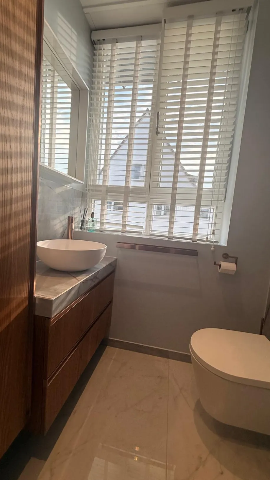 Schmales Badezimmer mit Holzwaschtisch, rundem Waschbecken und großem Fenster mit weißen Jalousien.