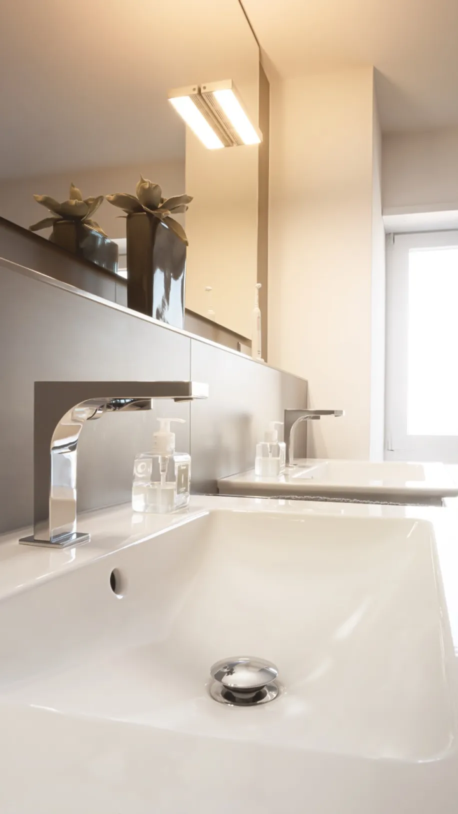 Detailaufnahme moderner Waschbecken mit verchromten Armaturen, großem Spiegel und heller Badezimmerbeleuchtung.