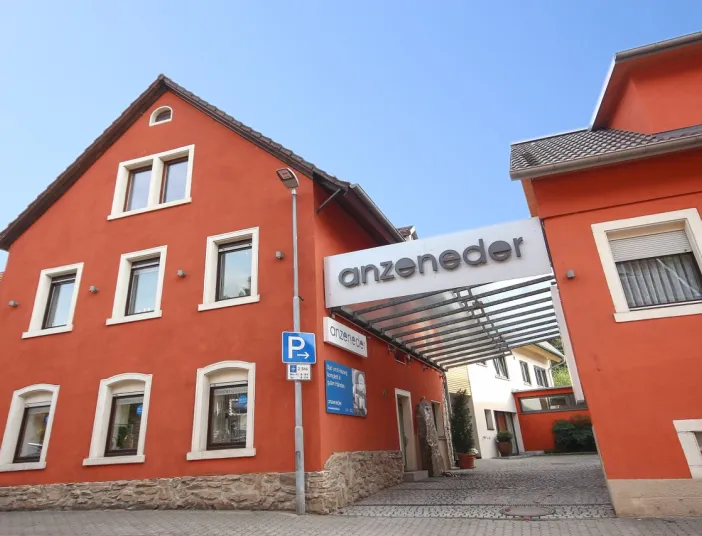 Außenansicht des Firmengebäudes von Anzeneder mit rot verputzter Fassade, überdachtem Eingangsbereich und Firmenlogo an der Straßenfront.